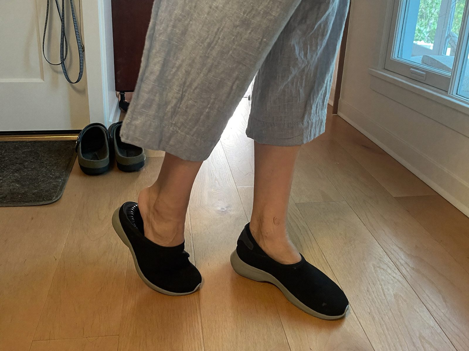 A woman in grey linen trousers stepping into a black knit Ample Daily sneaker in a hallway without using her hands